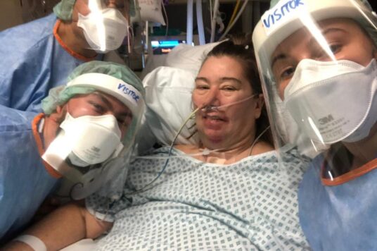 Jan in a hospital gown and lying on a bed with her family dressed n PPE