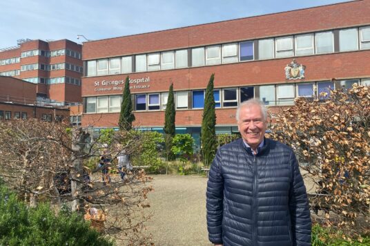 Martin Tyler wears a blue padded jacket and stands outside St George's Hospital
