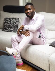 Dave sits on a sofa wearing a pink suit