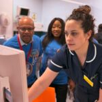 A staff member using the new electronic patient records system at a work station with two colleagues supervising