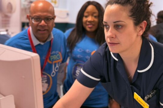 A staff member using the new electronic patient records system at a work station with two colleagues supervising